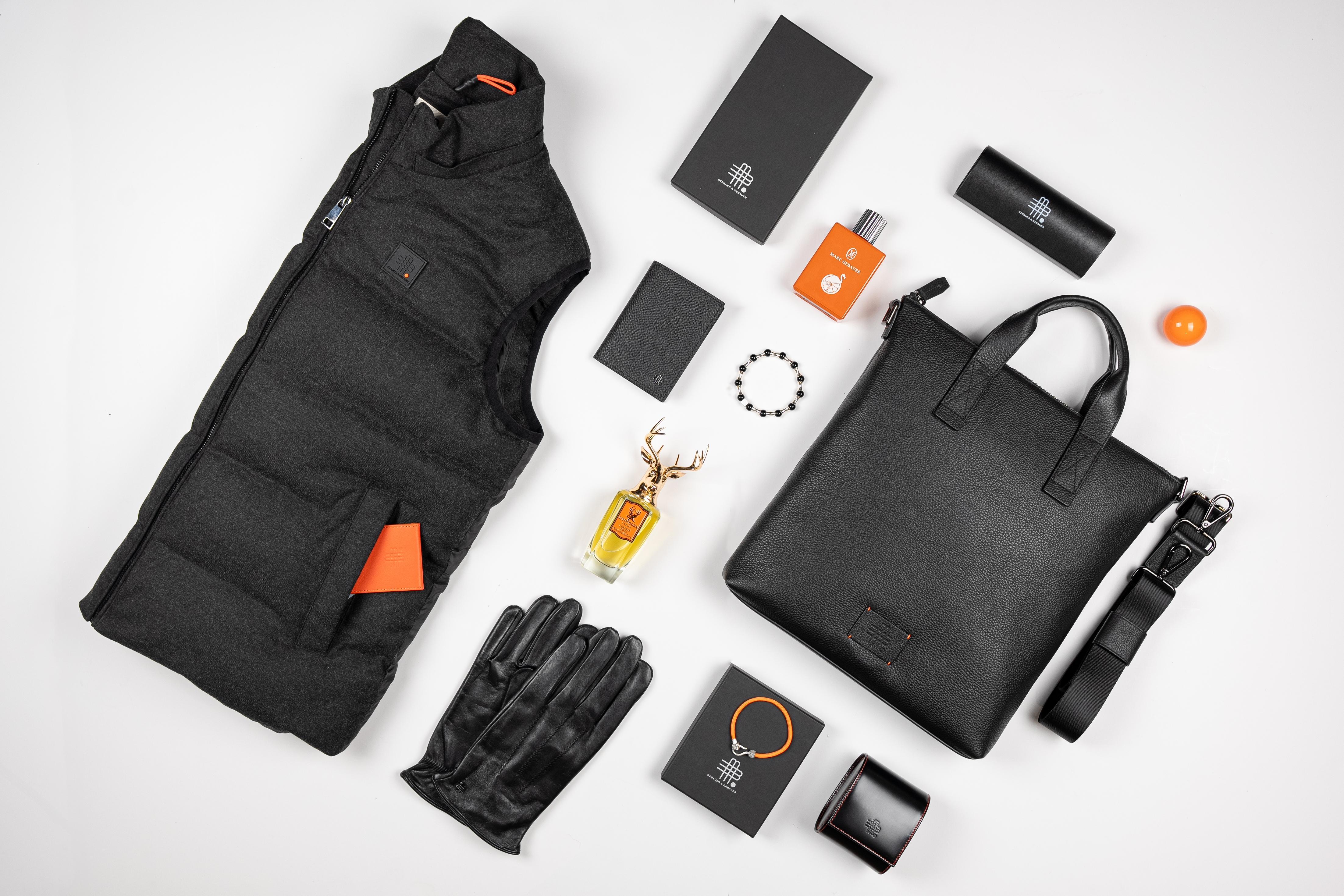 Gebauer und Gebauer Produktarrangement mit schwarzer Weste, Lederhandschuhen, schwarzer Ledertasche, Parfums, Gürtel und Accessoires auf hellem Hintergrund